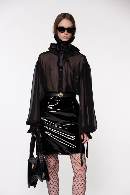 Model wearing the Helen Anthony black organza neck frill with detachable satin flower tie, styled with a sheer black silk organza shirt, PU-coated high shine leather skirt and small shoulder black leather bag