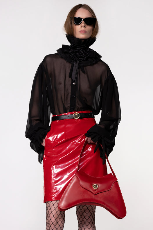 Model wearing the Helen Anthony black organza neck frill with detachable satin flower tie, styled with a sheer black silk organza shirt, PU-coated high shine leather skirt and small shoulder red leather bag
